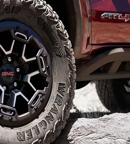 A close-up of an off-road Goodyear Wrangler tire on a black GMC wheel beside a red AT4X body panel, resting on a rocky surface.