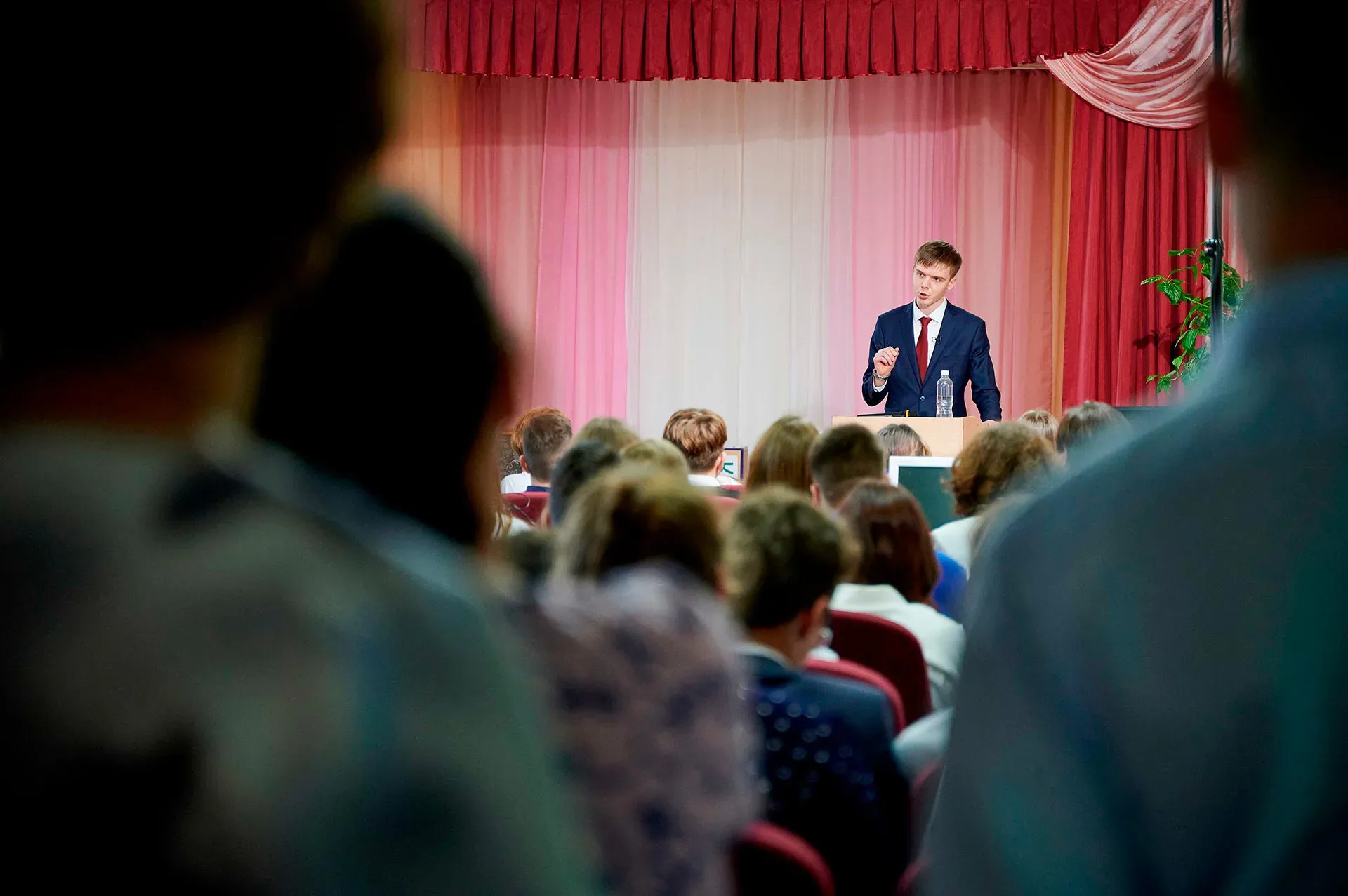 <Andrew Reutsky in a navy suit speaks from a podium to an audience seated in an auditorium with red curtains and pink drapery on the stage behind him.>