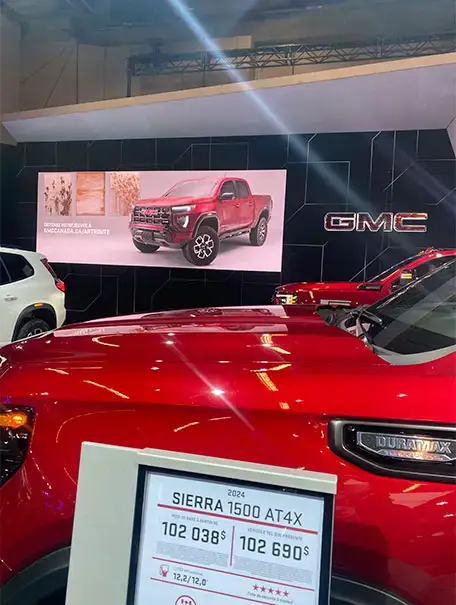 红色 GMC Sierra 1500 AT4X 在车展上展出，旁边有大型显示屏、价格牌以及其他 GMC 车型。