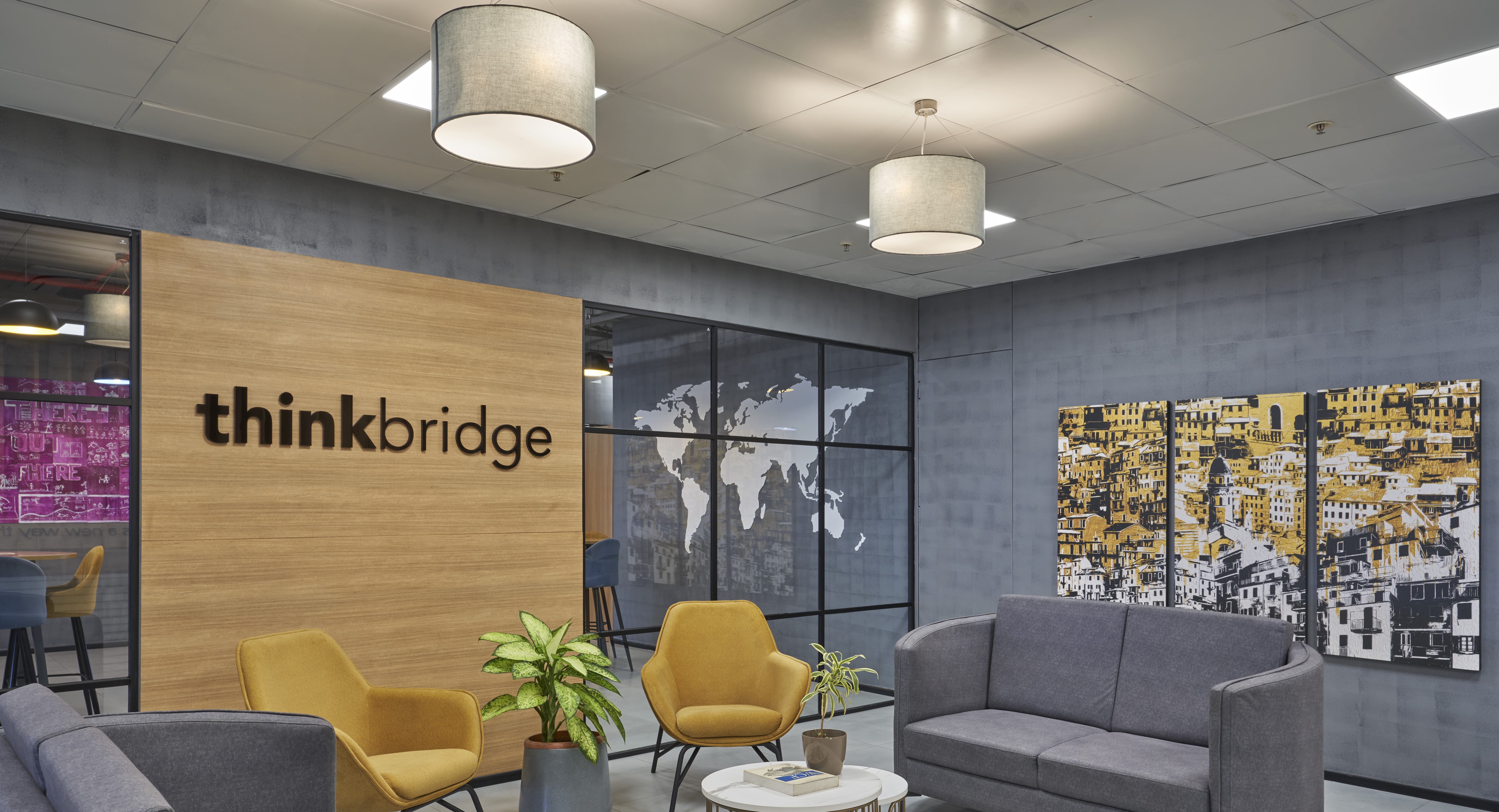 Modern office lounge with gray sofa, mustard chairs, plants, round white table, wooden wall with 'thinkbridge' logo, and triptych cityscape artwork.
