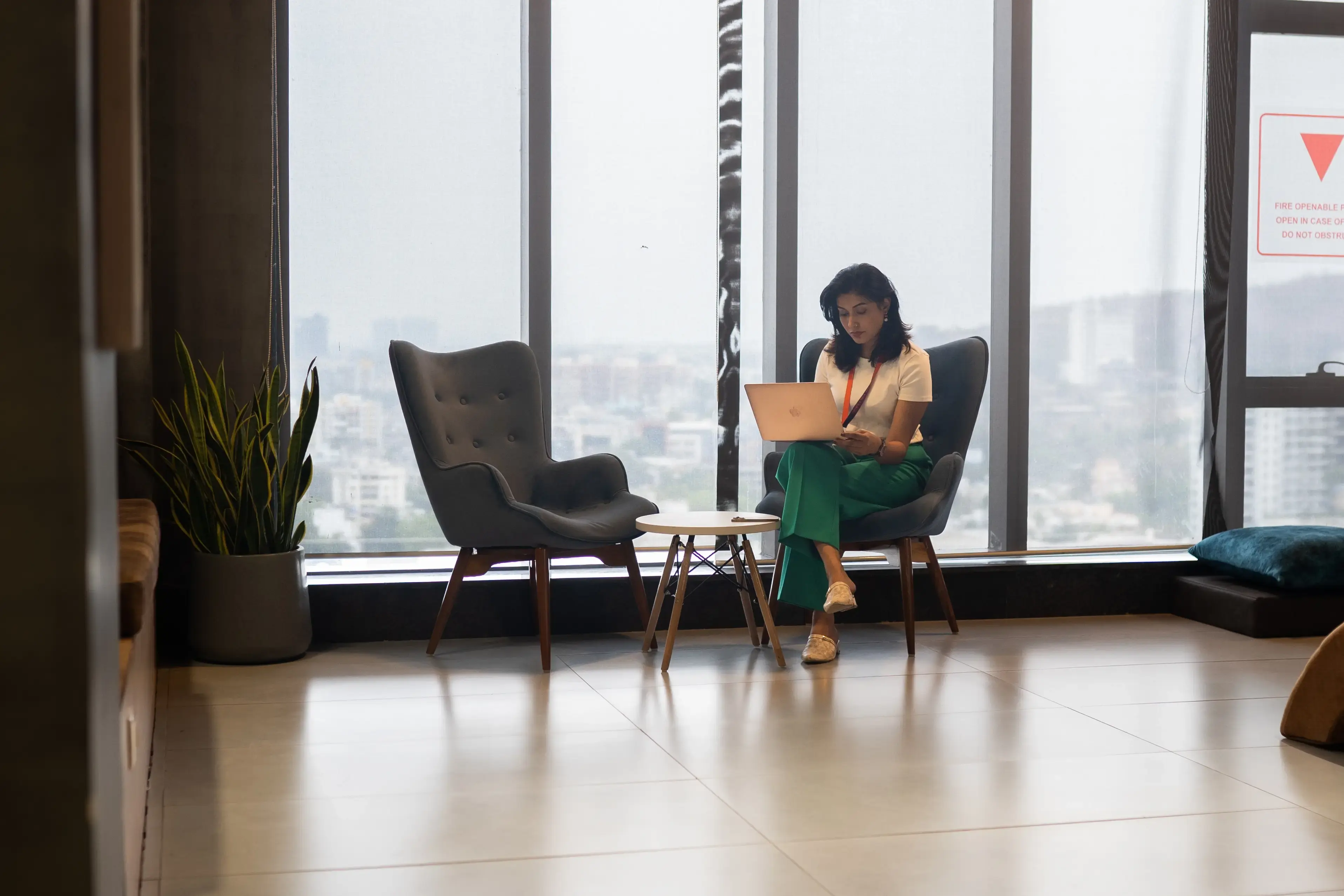 Woman sitting on a chair working on a laptop in a modern office with large windows and city view.