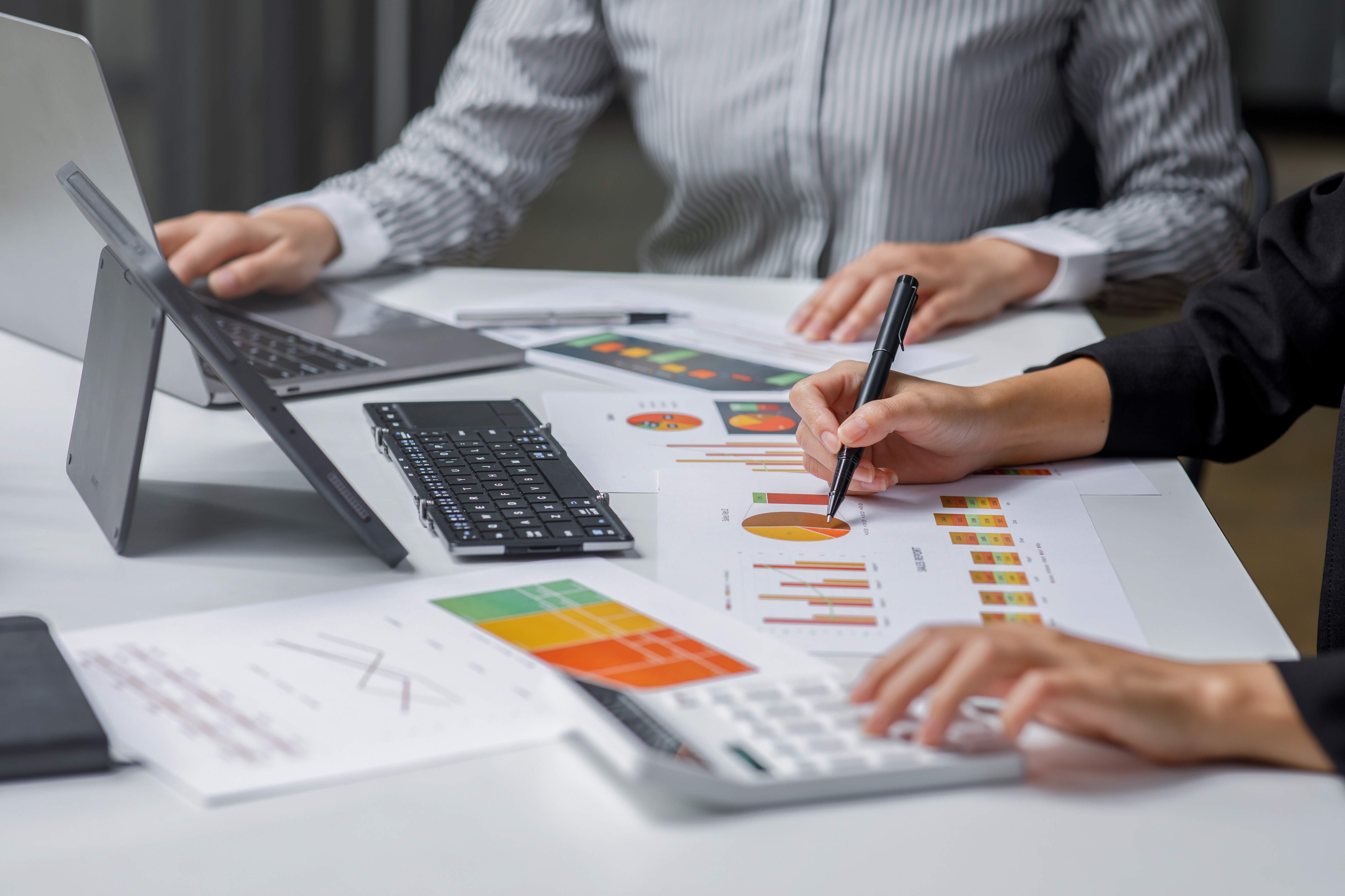 Two people working at a desk with financial charts, a calculator, a laptop, and a tablet keyboard, one person pointing at a pie chart with a pen.
