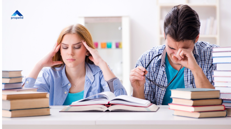 Two distressed students, a woman holding her temples and a man rubbing his eyes while holding glasses, sitting at a desk piled with textbooks.
