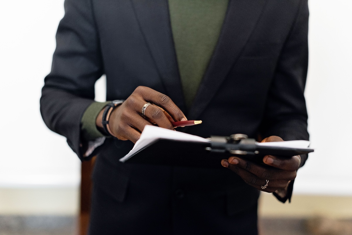 Close-up of an adult in a blazer holding a clipboard with documents and a pen indoors.