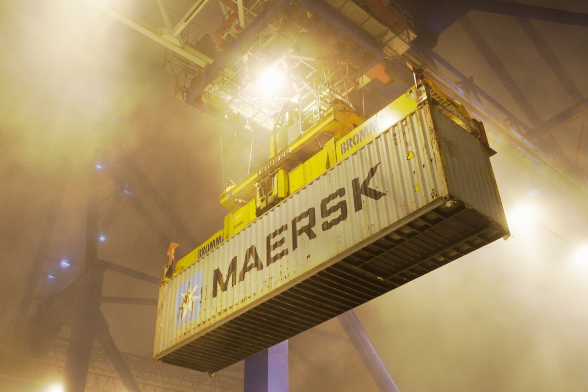 Industrial shipping container suspended by crane in Gdańsk port at night.