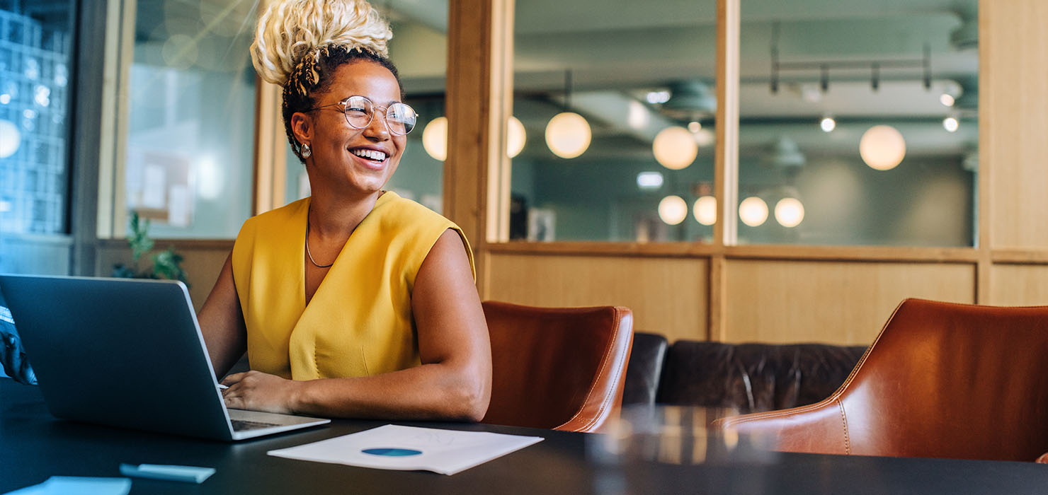 A diverse woman sitting in a leather office chair looks up from her computer and smiles.