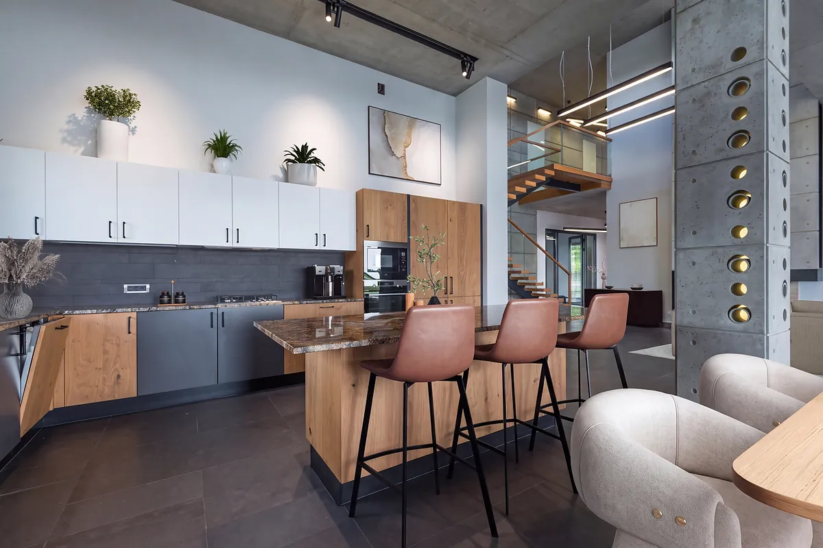 Loft kitchen staged by Apply Design: tan leather barstools at the granite island, framed artwork on the wall, potted plants along the upper cabinets, dried florals and a boucl&eacute; armchair in the foreground.