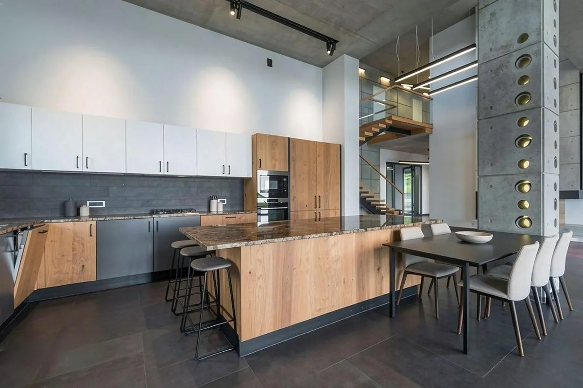 The same loft kitchen staged by REimagineHome: plain dark barstools at the island and a grey dining table with upholstered chairs to the right.