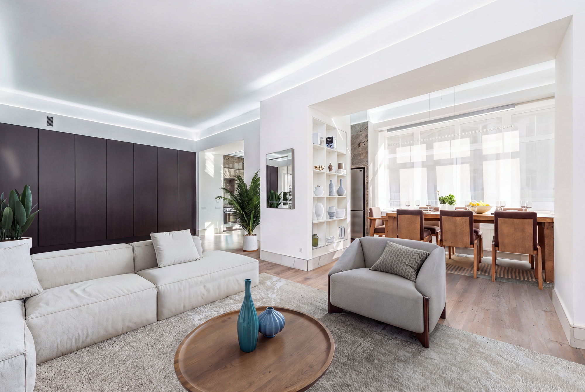 A third angle of the same apartment, staged with the sectional sofa, a round coffee table, an armchair and the dining set in the window bay