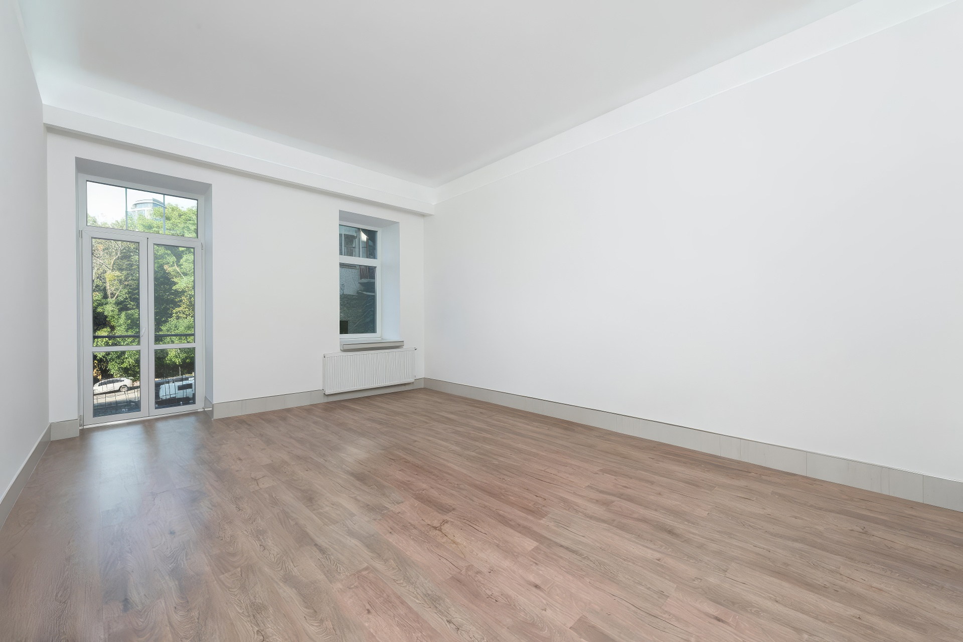Empty bedroom with pale wood flooring, a balcony door, a small second window and a radiator, photographed for a real estate listing