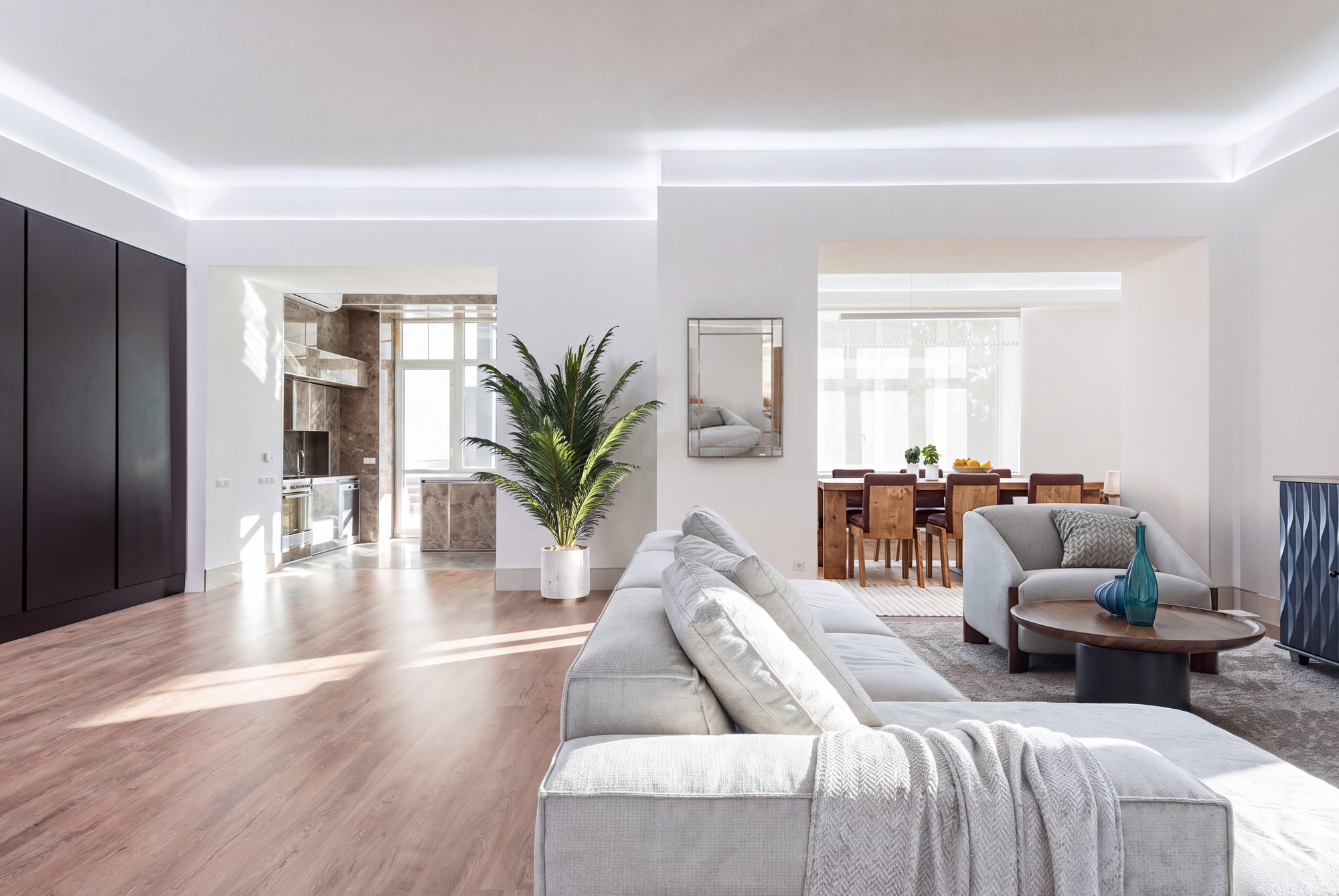 The same apartment from a second angle, staged with a grey sectional sofa, an armchair, a plant and the same dining set beyond