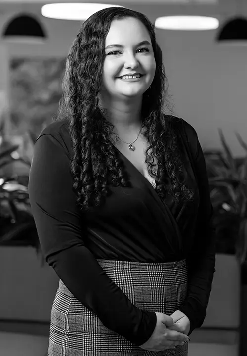 Smiling woman with curly hair wearing a black top and patterned skirt standing indoors.
