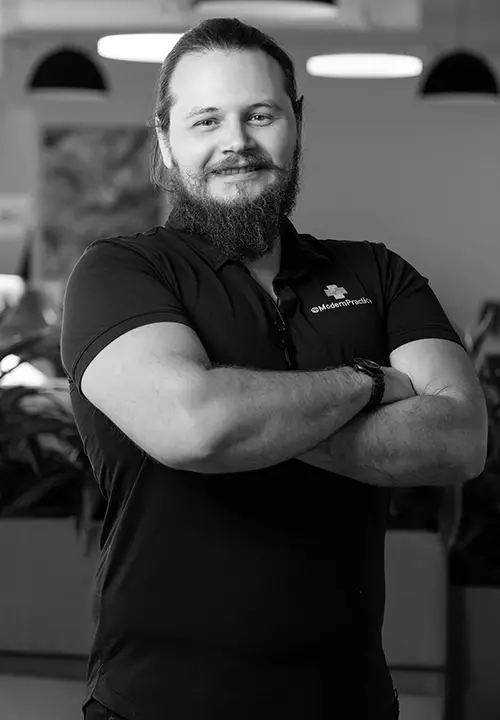 Smiling bearded man with crossed arms wearing a Modern Practice polo shirt in an office setting.