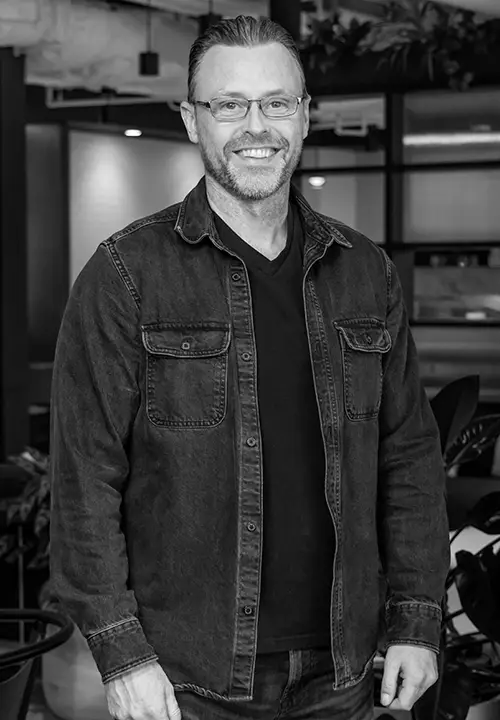 Smiling man with glasses in a black shirt and jacket standing in an indoor office setting.