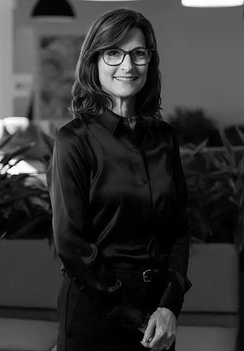Smiling woman wearing glasses and a black button-up shirt standing indoors with plants in the background.