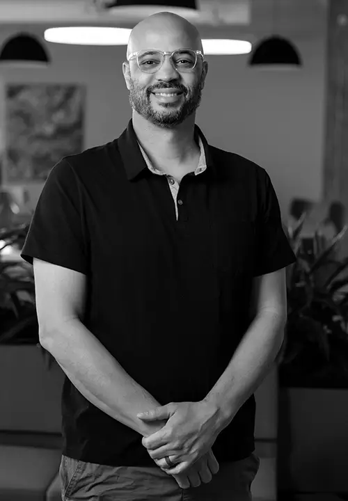 Smiling bald man with glasses in black polo shirt stands with hands clasped in an indoor setting.