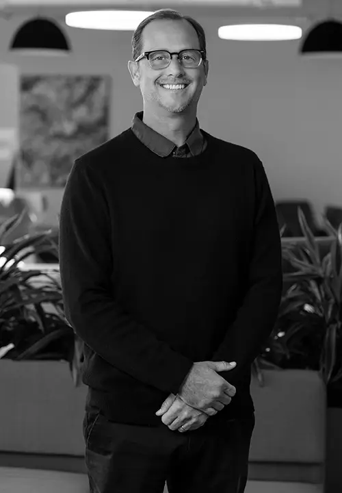 Smiling man with glasses and dark sweater standing indoors with plants and lights in the background.