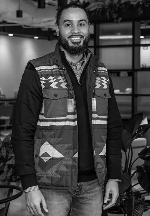 Smiling man with beard wearing patterned vest and black shirt standing indoors near plants.