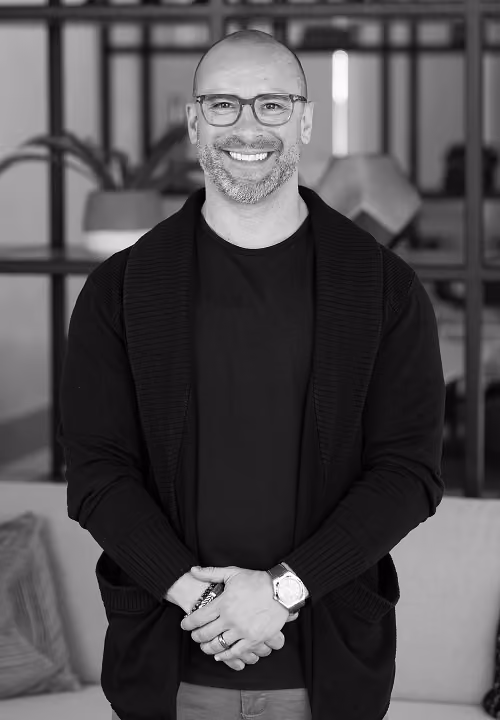 Founder, Brian Leffler, smiling wearing glasses and a black polo shirt standing with hands clasped indoors.