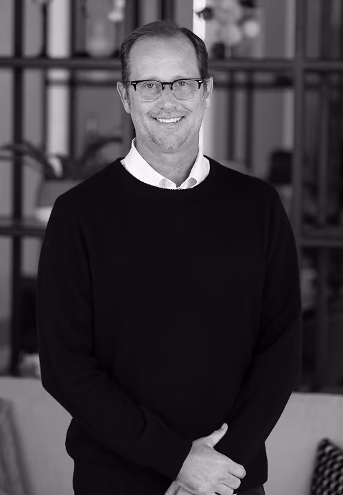 Smiling man with glasses and dark sweater standing indoors with plants and lights in the background.