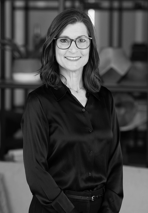 Smiling woman wearing glasses and a black button-up shirt standing indoors with plants in the background.