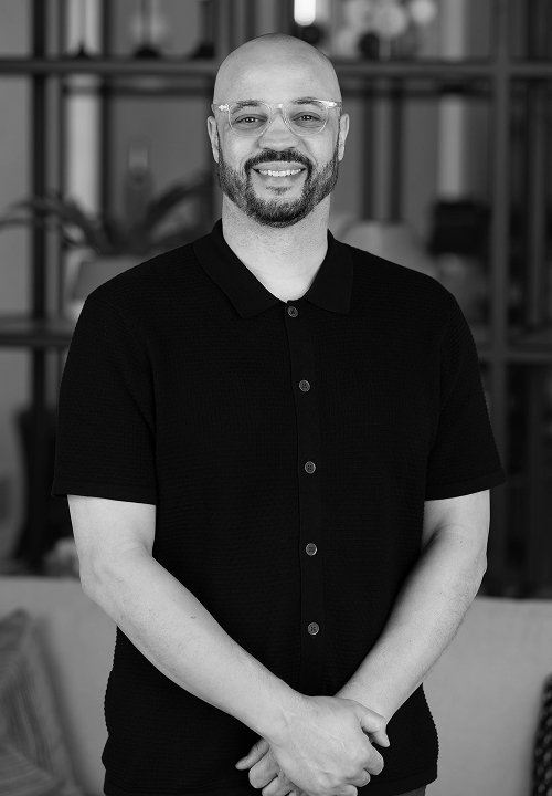 Smiling bald man with glasses in black polo shirt stands with hands clasped in an indoor setting.