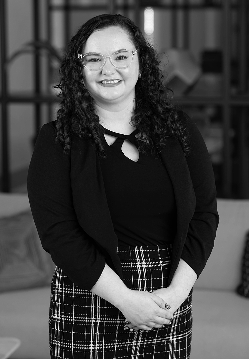 Smiling woman with curly hair wearing a black top and patterned skirt standing indoors.
