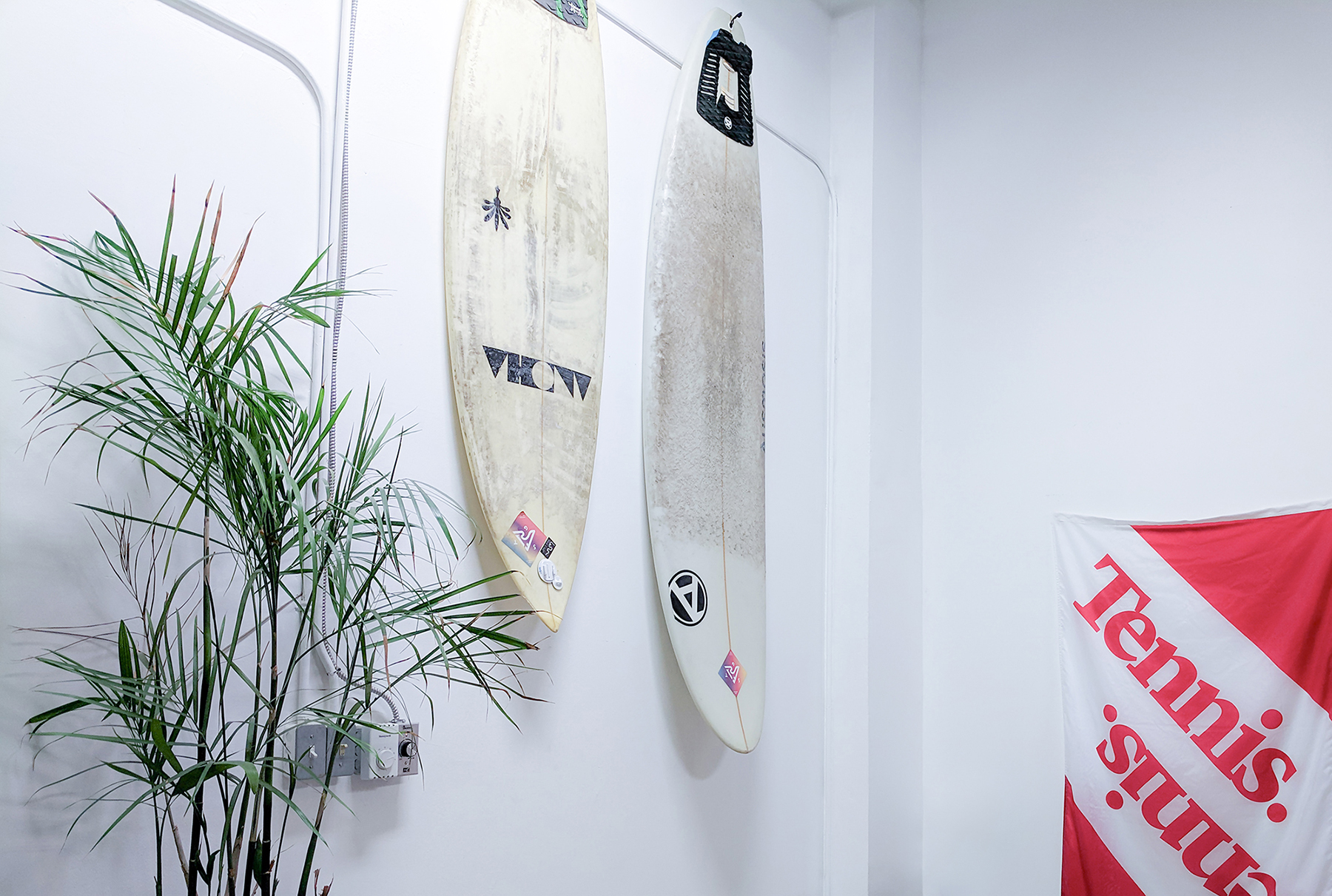 Against the starkness of a white wall, two surfboards stand sentinel, their once-gleaming surfaces telling tales of sun-soaked adventures with a patina of salt and sea. A potted palm, green and vibrant, whispers of tropical breezes in the quiet corner it occupies. To their side, a bold flag, emblazoned with the words 'Tennis' flutters in still air, a vibrant splash of red and white proclaiming a passion for the game. Together, they compose a silent ode to leisure and the call of the ocean waves.