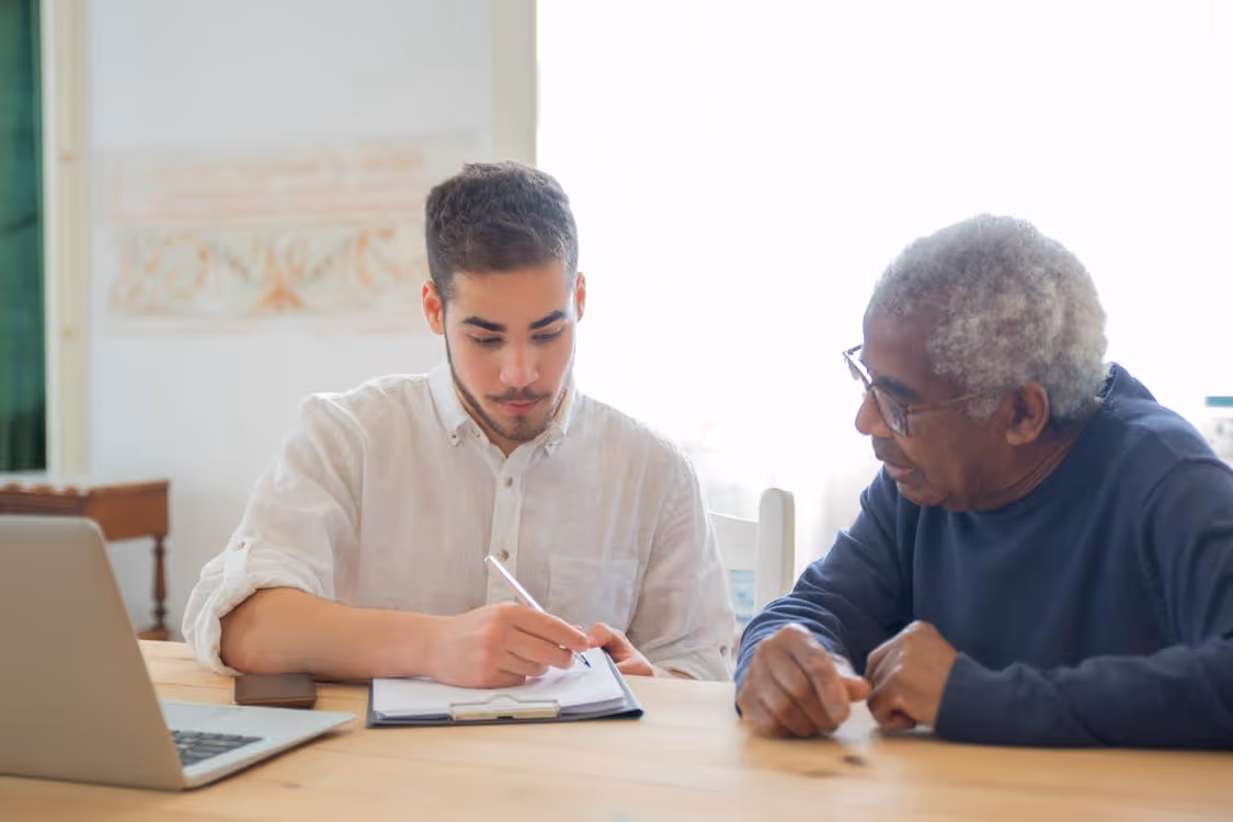 Free Young man helps elderly man with documents at a desk indoors, promoting collaboration. Stock Photo