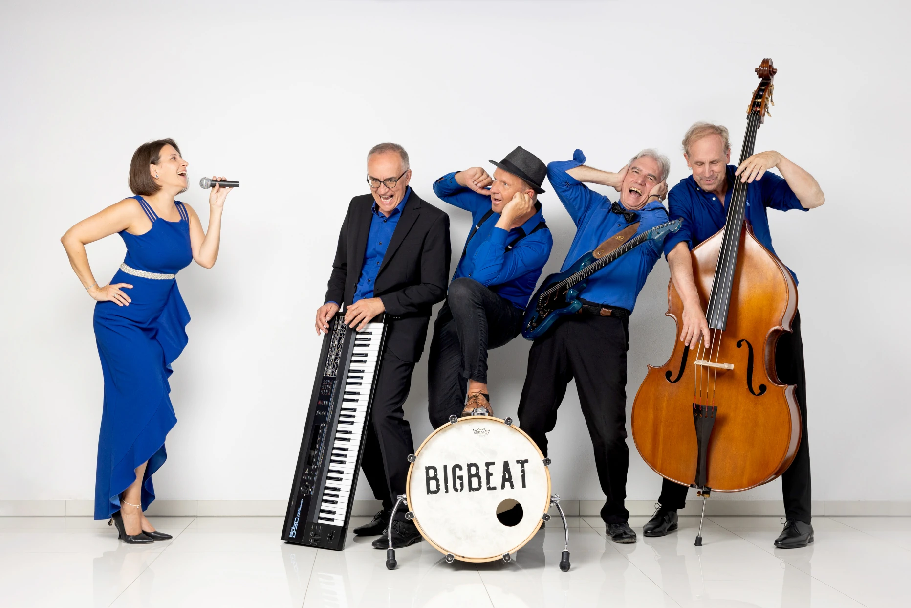 BigBeat – Event- und Hochzeitsband: Fünf Musiker in blauen Outfits posieren humorvoll mit Sängerin, Keyboard, Gitarre, Kontrabass und Bassdrum vor weißem Hintergrund.