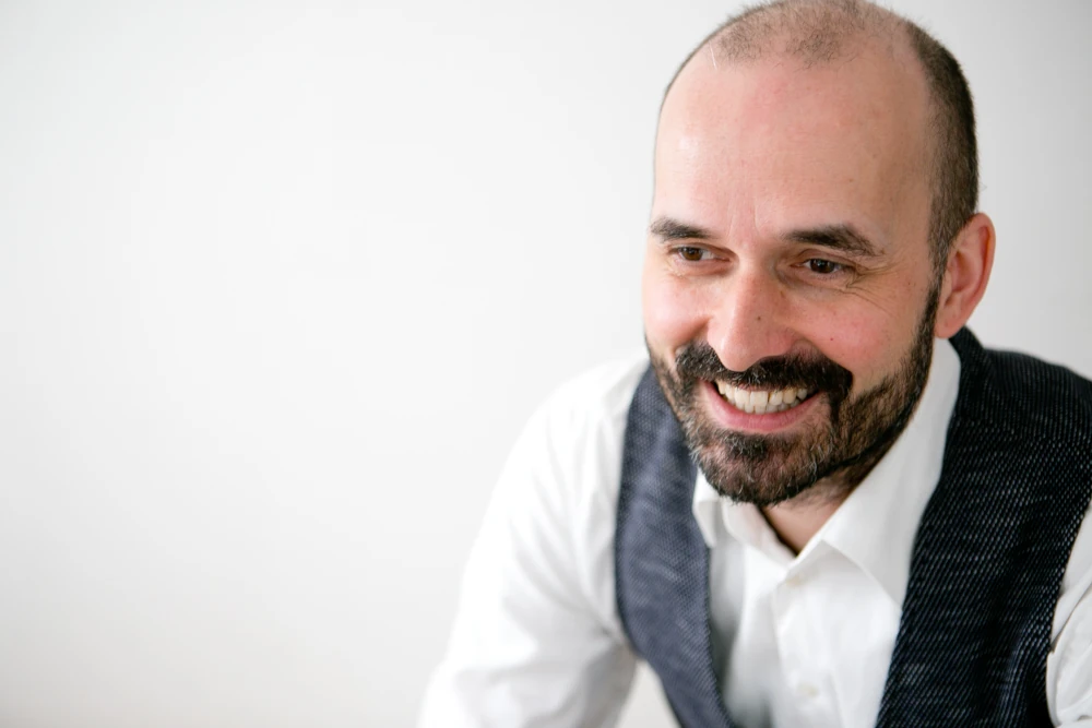 Alexander Dressler, ein lächelnder, bärtiger Mann mit Glatze, weißem Hemd und grauer Weste vor weißem Hintergrund.