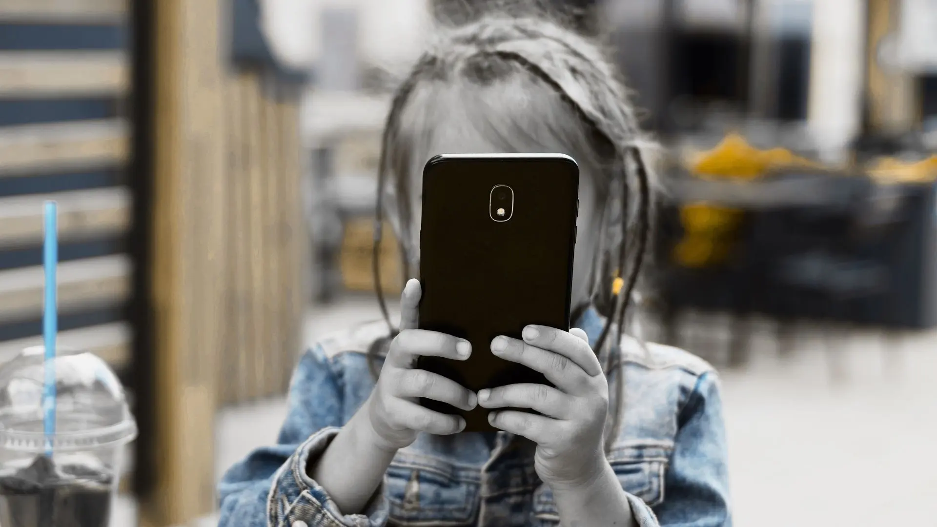 Fotoğrafta bir kız çocuğu elinde telefon tutuyor.