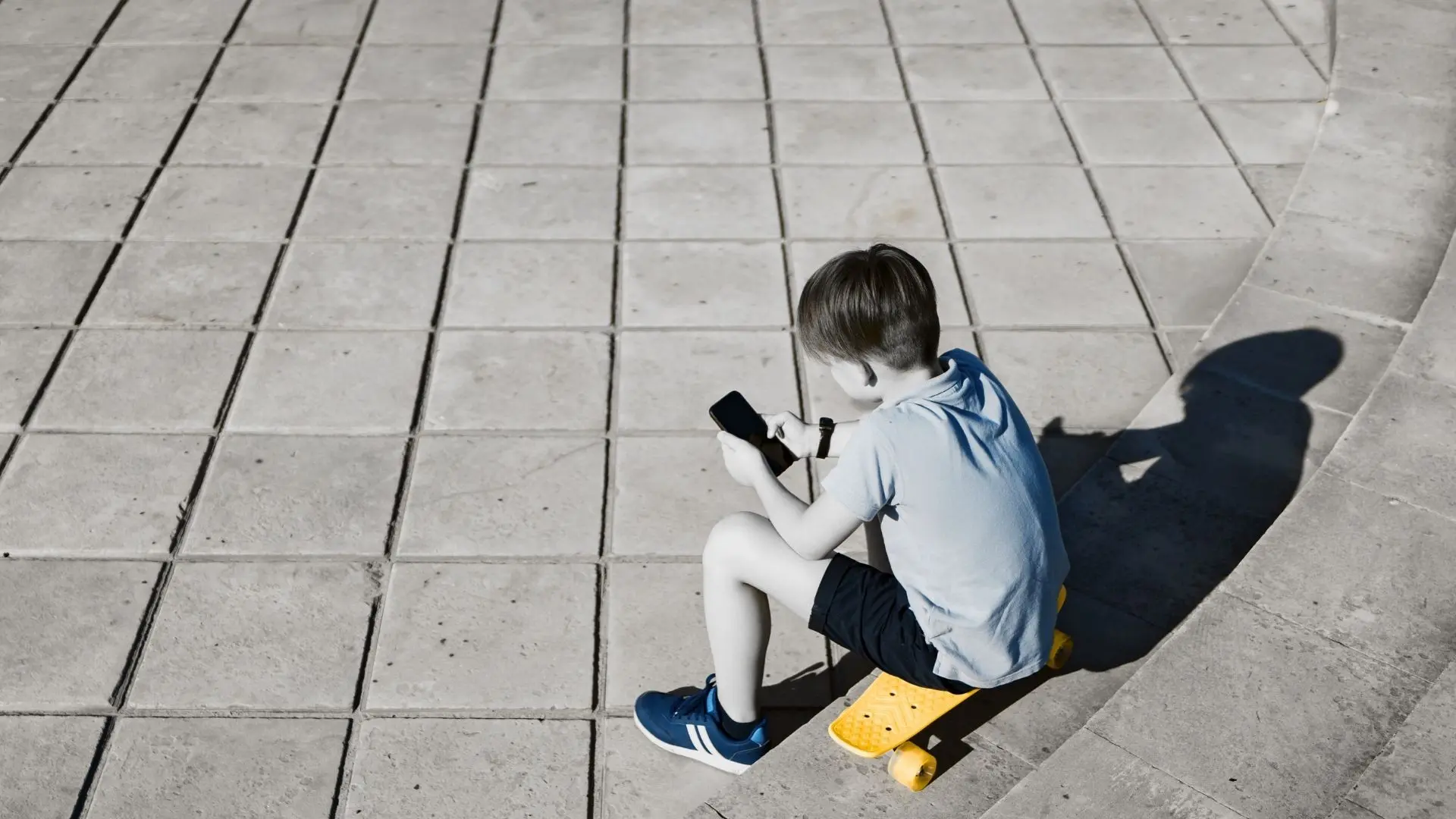 Fotoğrafta kaykay üstüne binmiş bir çocuk elinde telefonuyla oynuyor.