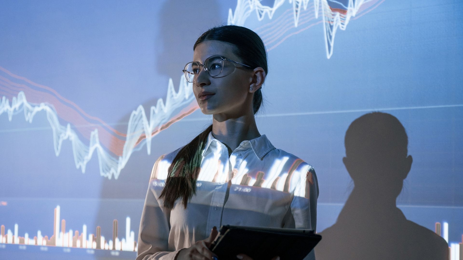 Kreatif stok görsel, gözlüklü topuz saçlı çalışan görünümlü bir kadının veri grafiği önünde duruşu gözüküyor.