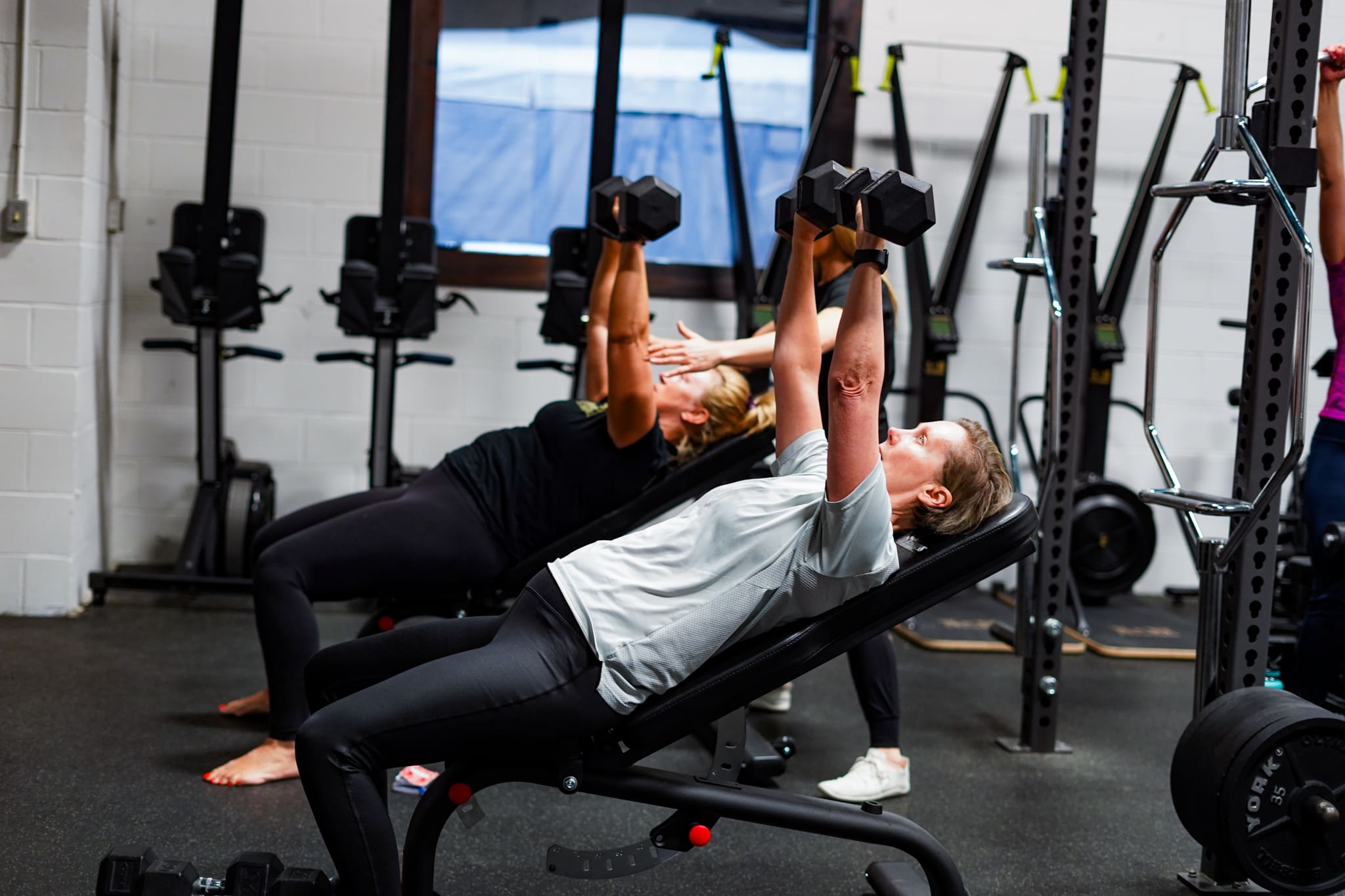 Two women lift a weight above their head. They low calm and controlled in the Move Strong 40+ programming.