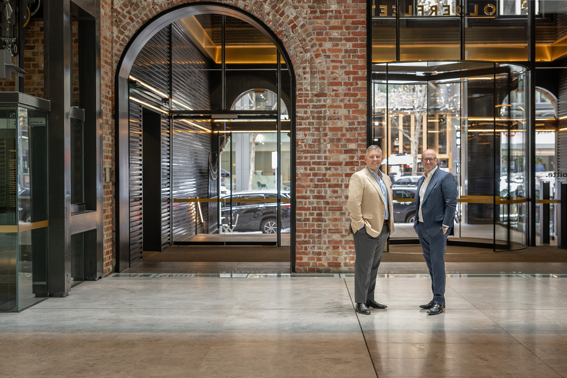 Two men in business attire standing and smiling in a spacious modern lobby with brick walls and large glass entrance doors.