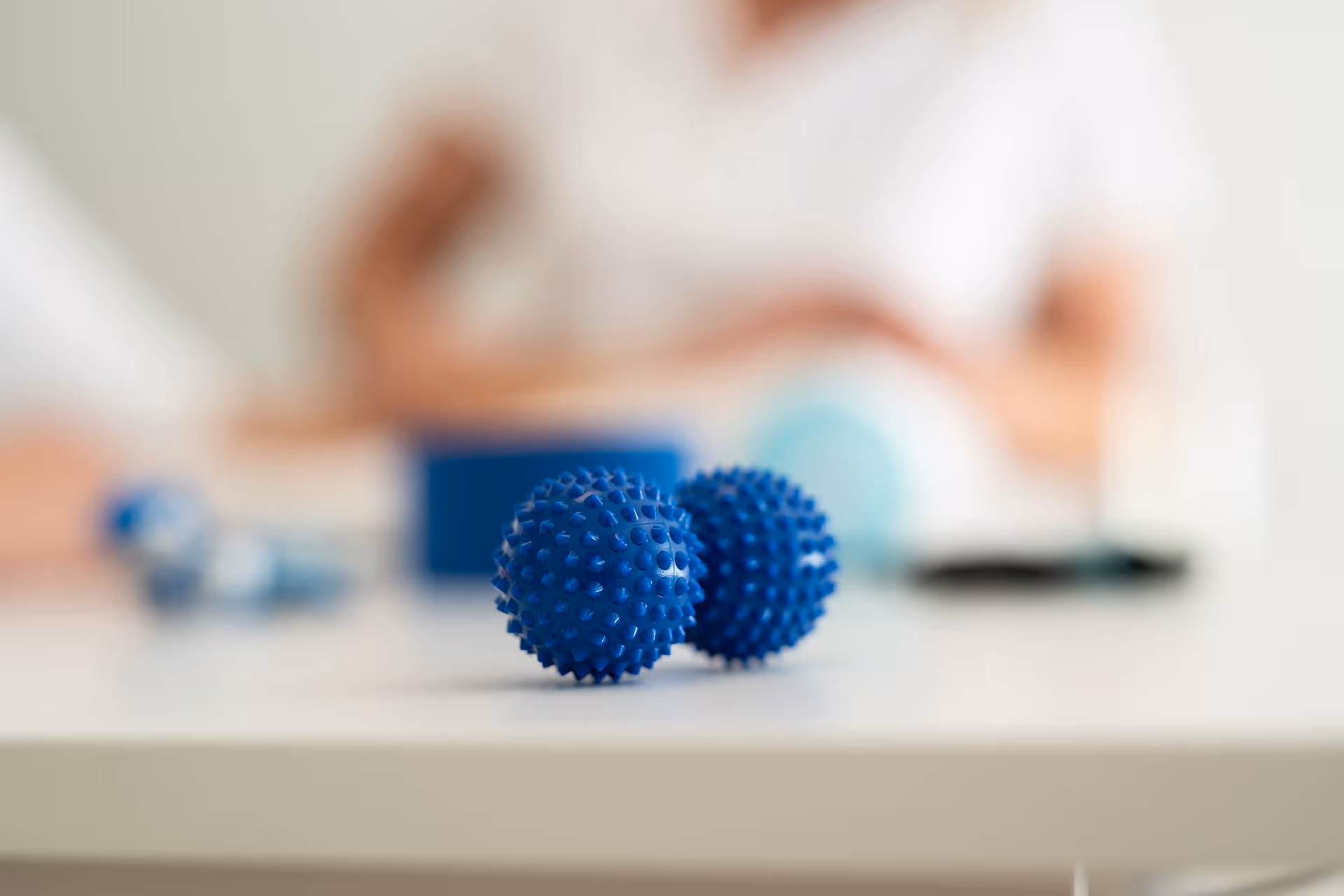 Zwei blaue Igelbälle für Hand- und Ergotherapie auf einem Tisch, im Hintergrund unscharf eine Therapeutin.