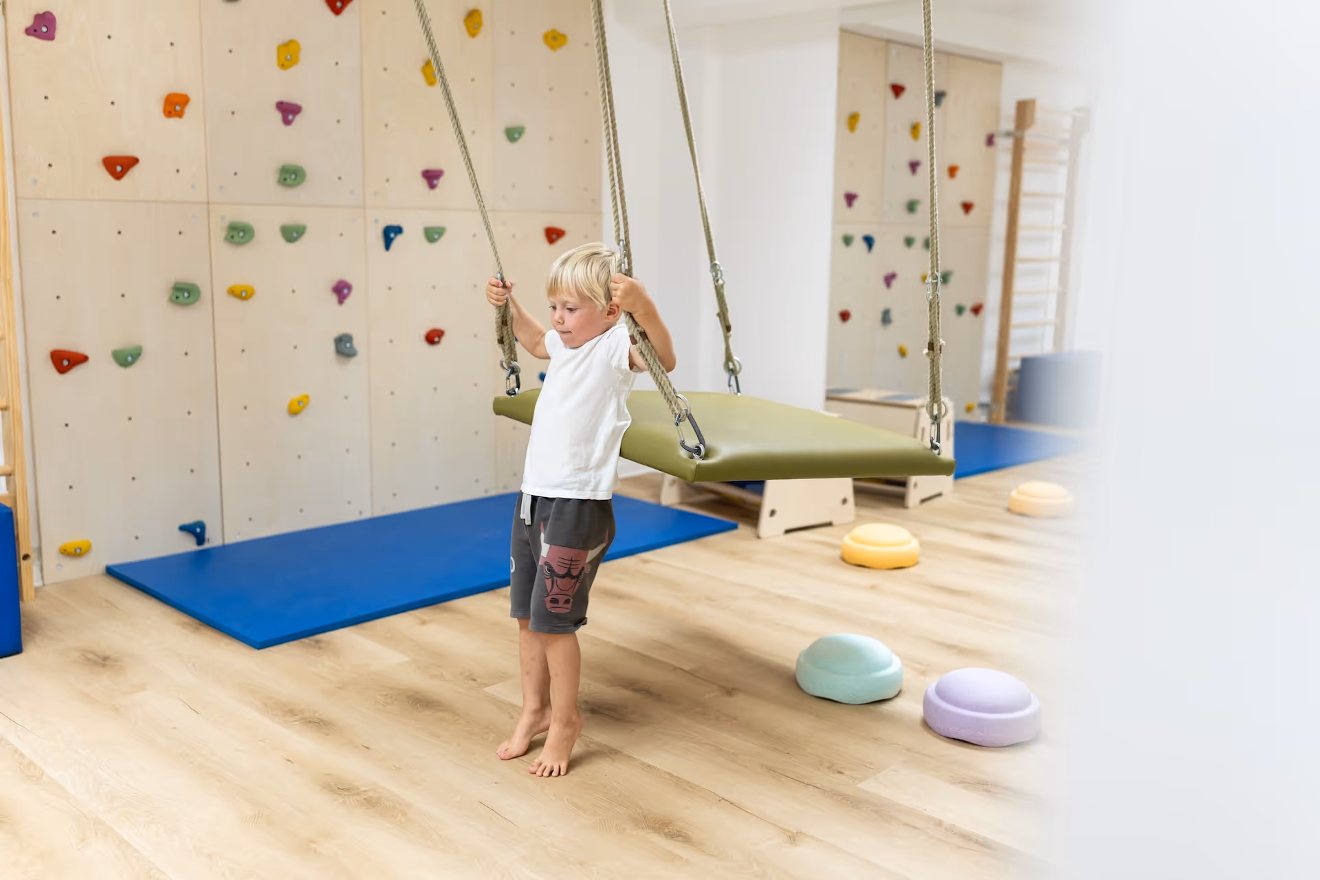 Blonder Junge mit weißem T-Shirt steht vor einer Brettschaukel in einem Therapieraum mit blauer Matte und Kletterwand.