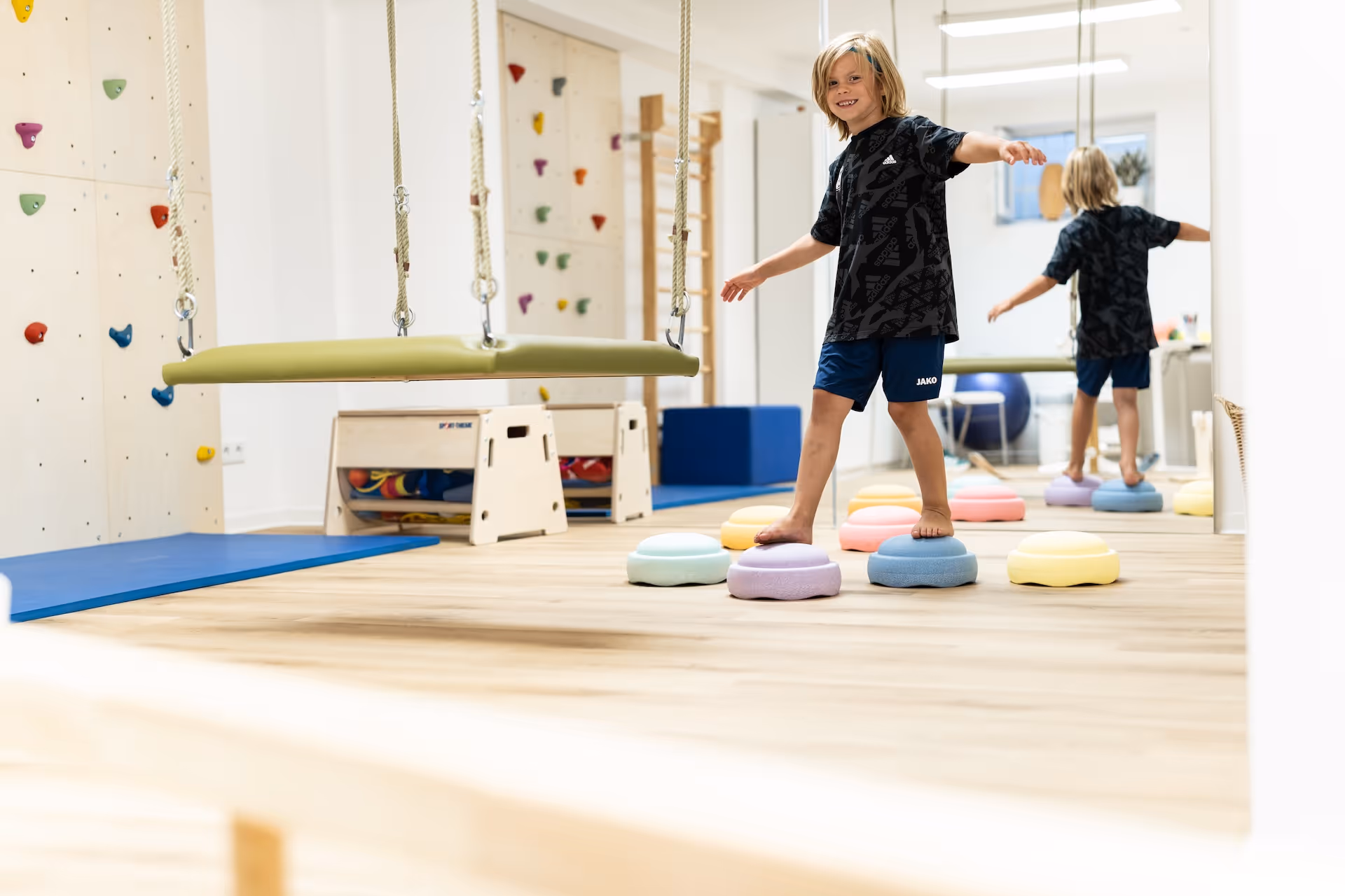 Junge steht mit ausgebreiteten Armen auf bunten Balacier-Steinen in einem Therapieraum mit Matten, Schaukel und Kletterwand.