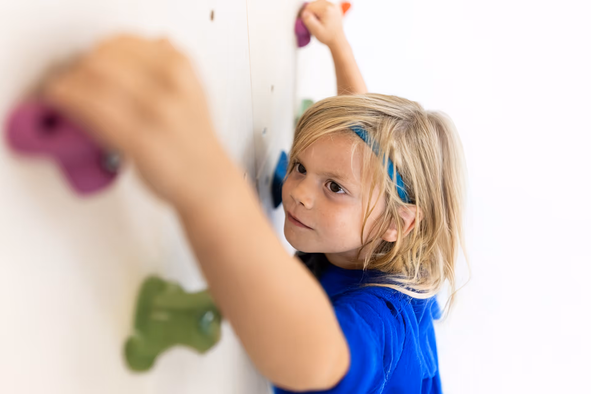Grinsender Junge blauem Shirt und Stirnband hält sich an einer Kletterwand mit roten und grünen Griffen fest.