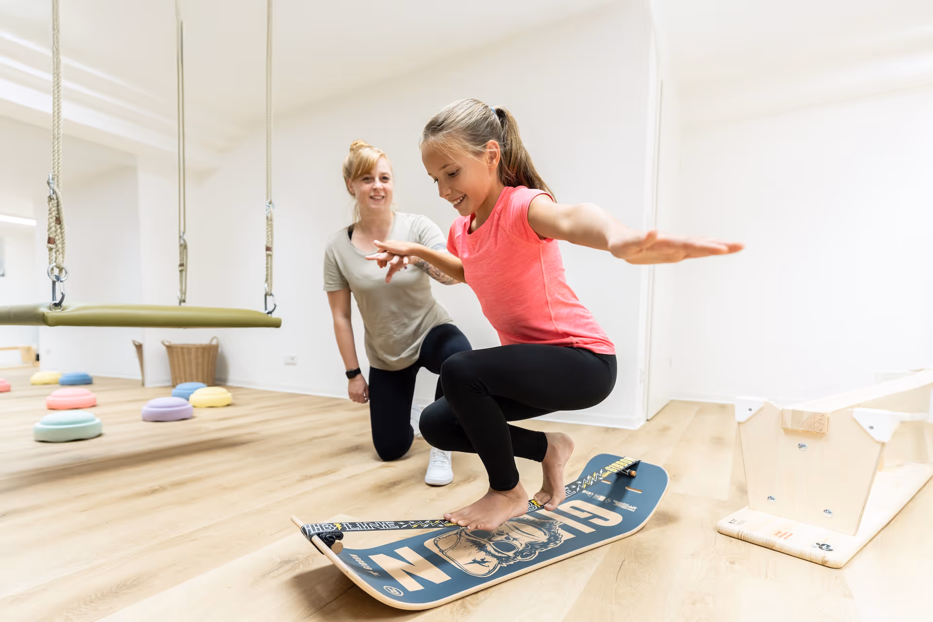 Lächelndes Mädchen mit pinkem Shirt hockt auf einem Balancier-Board und wird von einer knienden Therapeutin gestützt.