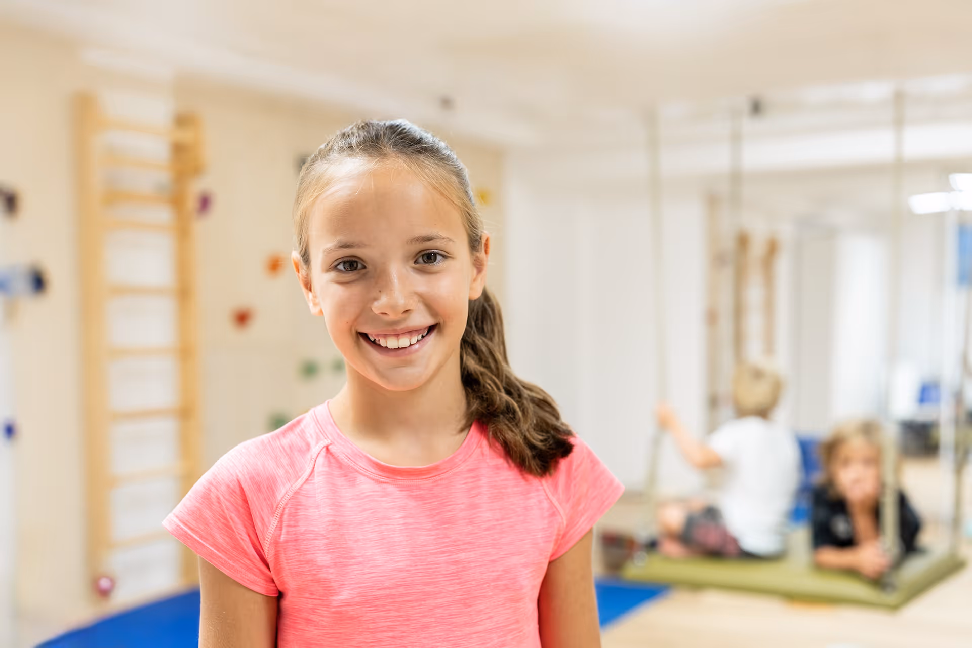 Lächelndes Mädchen in Sportkleidung steht in einem Therapieraum mit Kletterwand und Schaukeln