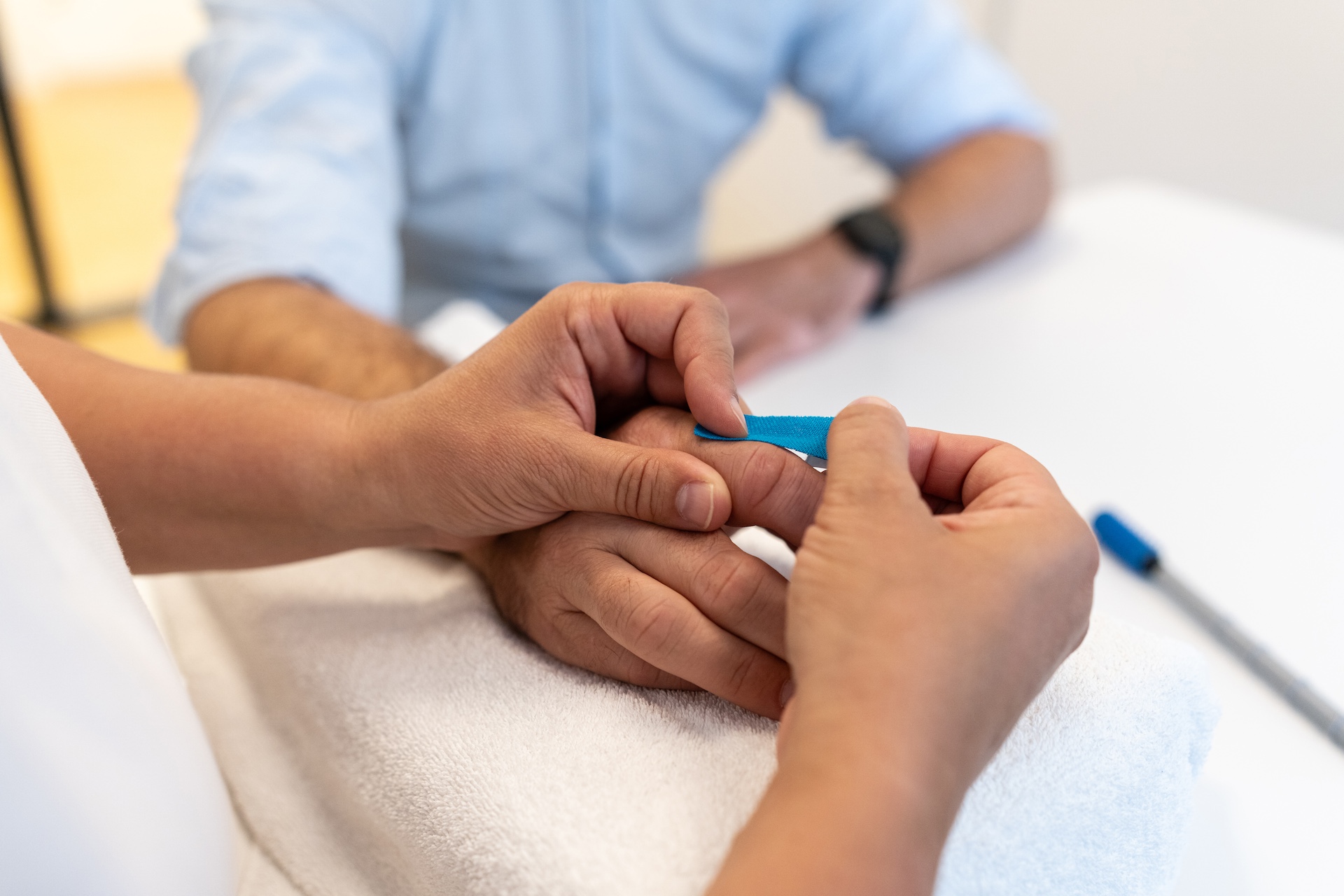 Therapeutin legt einer Patientin ein blaues Therapieband am Handgelenk an, beide tragen Schutzmasken.