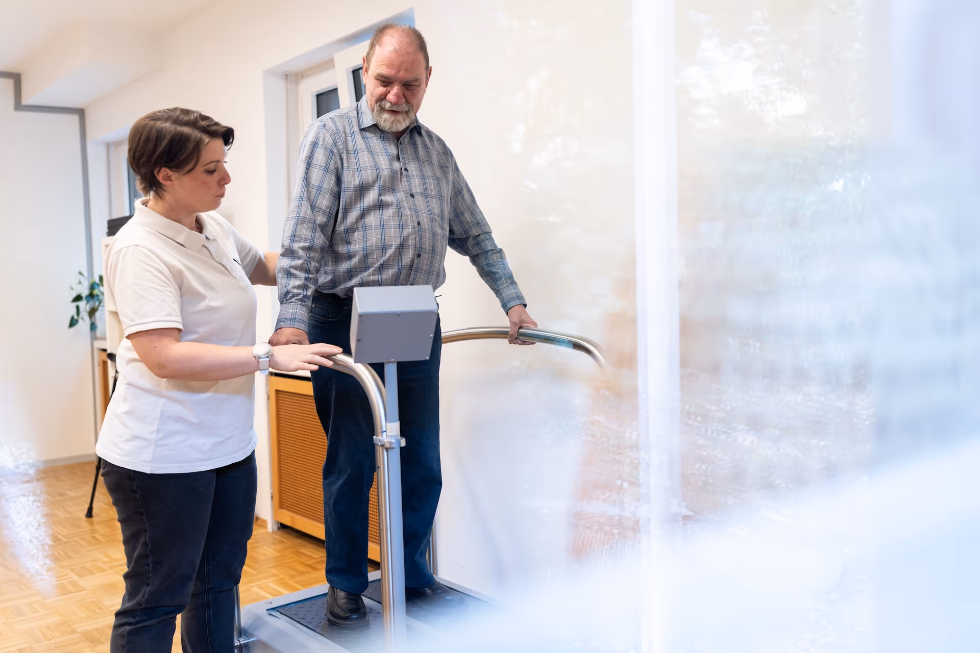 Therapeutin unterstützt einen älteren Mann beim Training auf einem Trainingsgerät mit Haltegriffen in der Praxis.