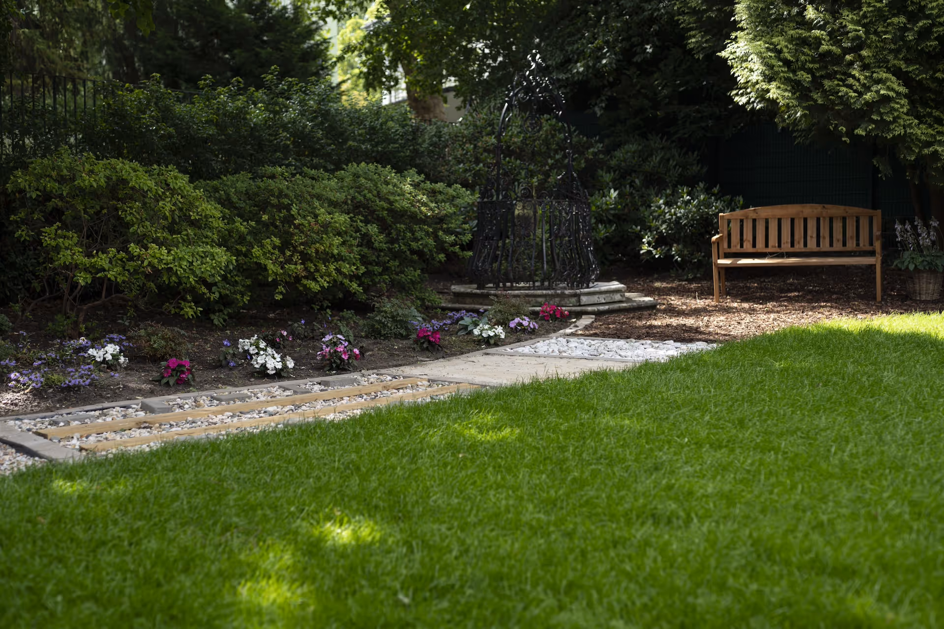 Gepflegter Garten mit grüner Rasenfläche, Blumenbeet und einer Holzbank im Schatten, umgeben von Sträuchern und Bäumen.