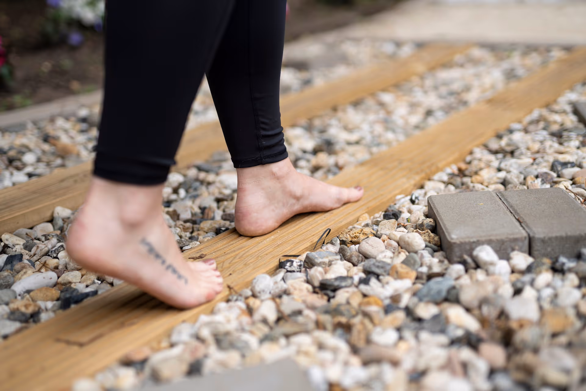 Person geht barfuß auf einem Weg aus Holzplanken und Kiessteinen.