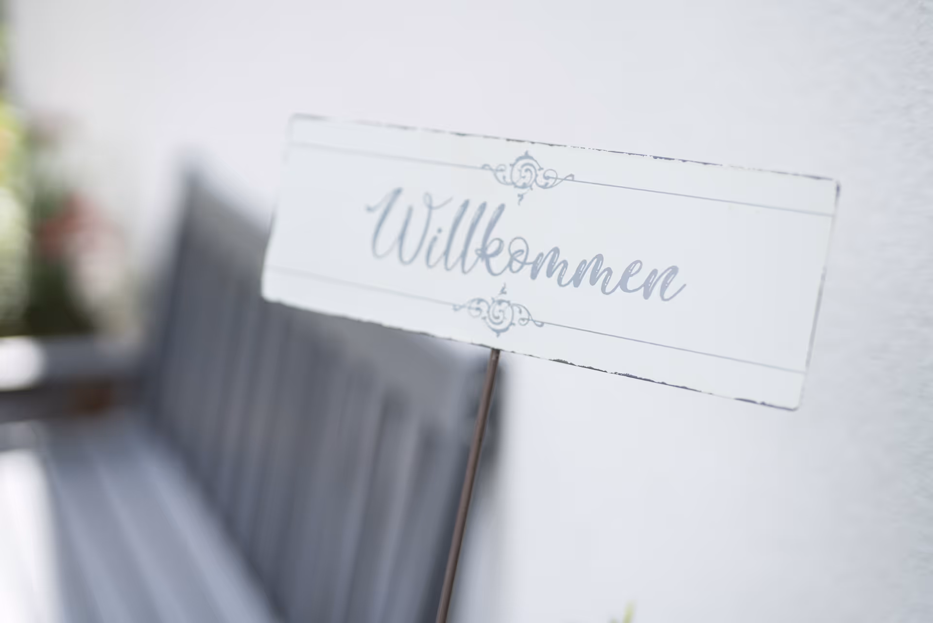 Willkommensschild vor einer weißen Wand mit grauer Sitzbank im Hintergrund.
