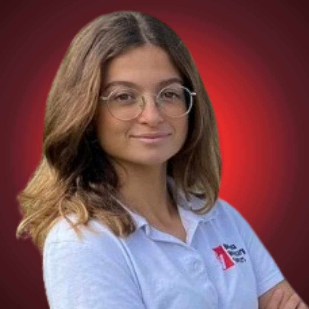 Young woman with shoulder-length brown hair and glasses, wearing a white polo shirt with a red and black logo, standing against a dark red background.