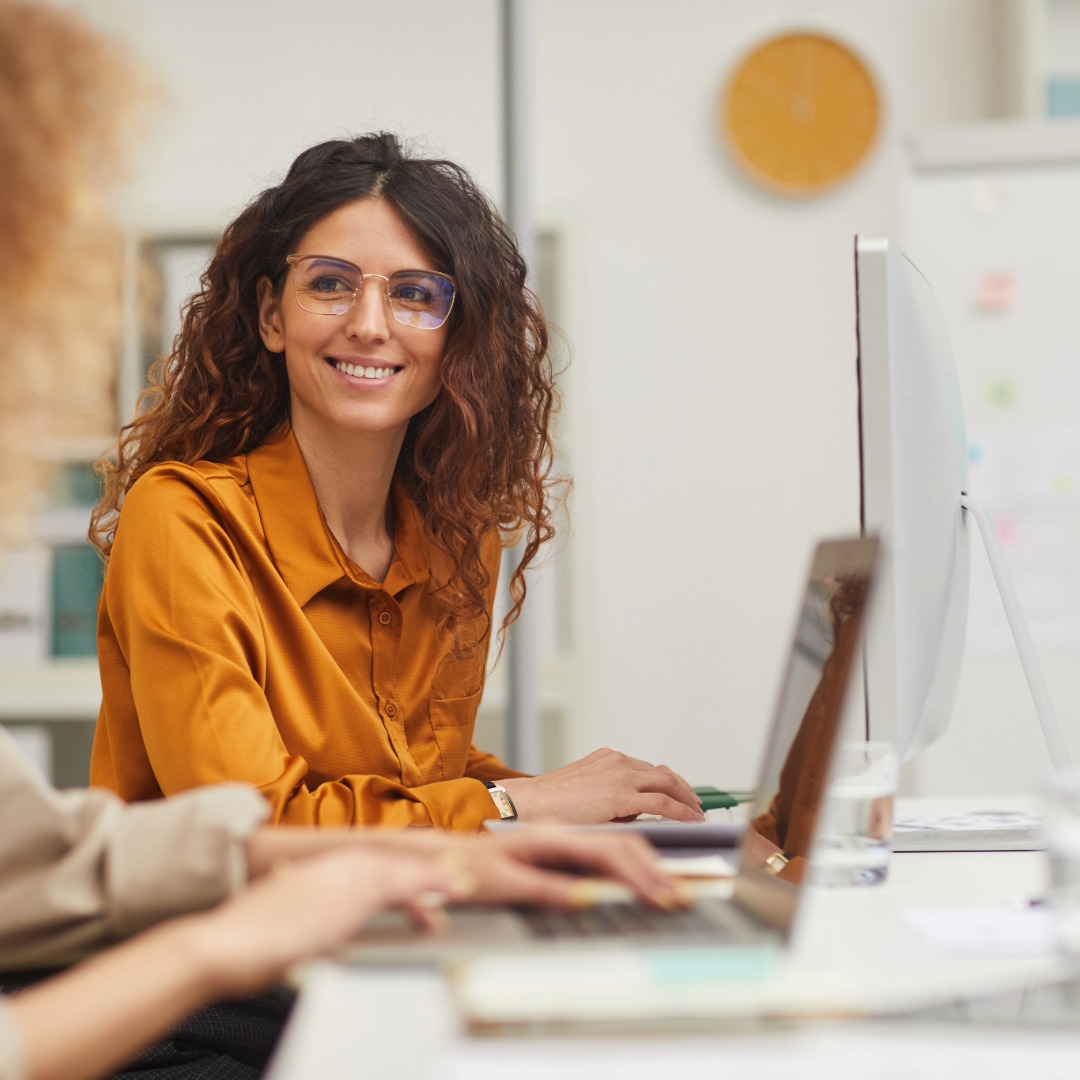 Smiling woman with curly hair and glasses wearing a mustard-colored shirt, sitting at a desk working on a computer in an office.