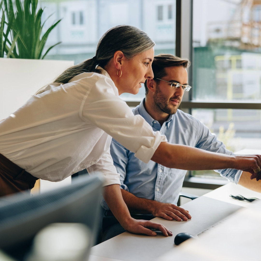 Woman leaning over to assist a seated man at a desk with a computer in an office.